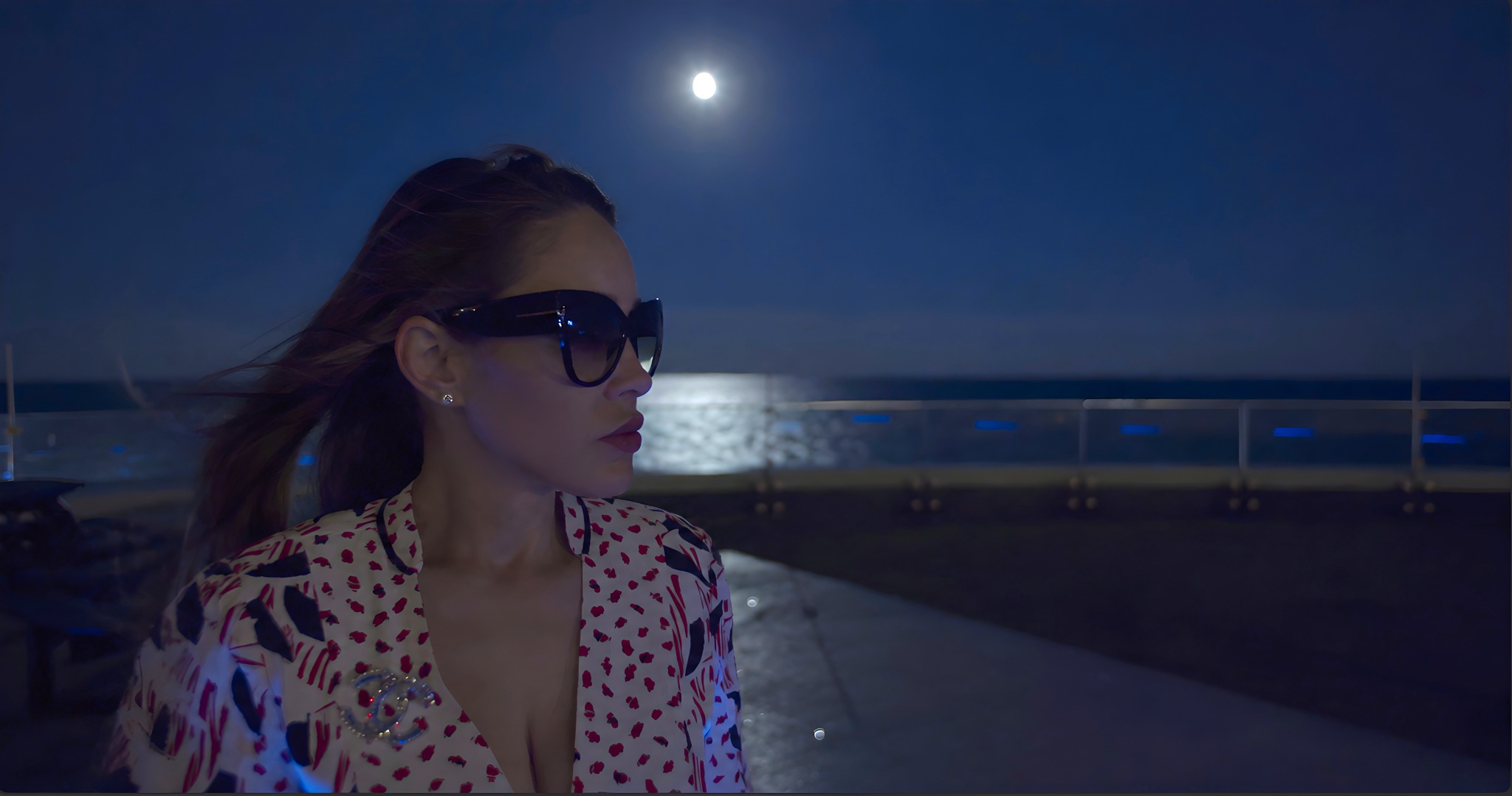 A woman in sunglasses beside the ocean under a silver moon, in deep midnight blue light.